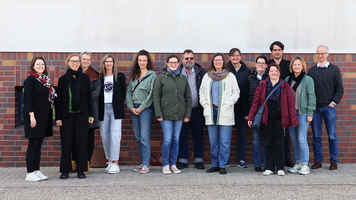 Gruppenfoto von 15 Personen, die in einer Reihe vor einer rot-braun geklinkerten Wand stehen. Alle blicken in die Kamera und lächeln. Die Kleidung ist lässig bis halbformell, die Stimmung freundlich und offen. Es handelt sich um Teilnehmende eines Zertifizierungsseminars.