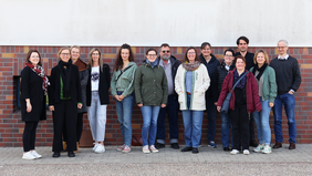 Gruppenfoto von 15 Personen, die in einer Reihe vor einer rot-braun geklinkerten Wand stehen. Alle blicken in die Kamera und lächeln. Die Kleidung ist lässig bis halbformell, die Stimmung freundlich und offen. Es handelt sich um Teilnehmende eines Zertifizierungsseminars.