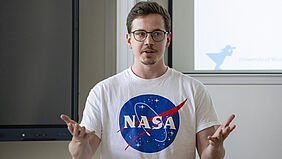 A young man wearing glasses and a white T-shirt stands in front of the projection screen for his PowerPoint presentation. He faces the audience with his arms open.