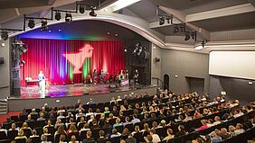 Der Blick geht von der Empore des Theaters über das Publikum Richtung Bühne. Dort ist an den roten Bühnenvorhang das Fischger-Signet aus dem Hochschul-Logo projiziert.