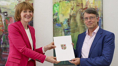 Die beiden Personen stehen vor einer Wand mit zwei großformatigen Bildern. Sie halten gemeinsam die Ernennungsurkunde in der Hand. 
