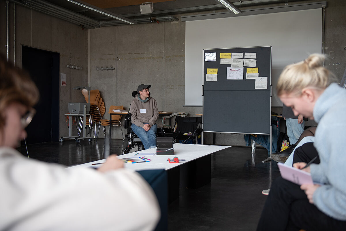 Mehrere Personen sitzen im Workshop und machen sich Notizen, während vorne eine Person neben einer Tafel sitzt und Inhalte erklärt.