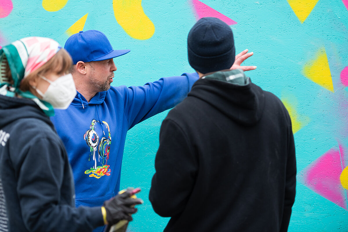 Scraby, ein Graffiti-Künstler, demonstriert Sprühtechniken an einer Wand, während Teilnehmende des Workshops zusehen.