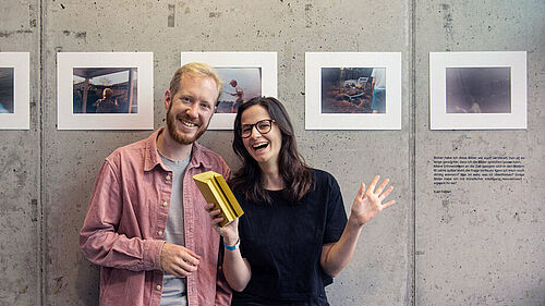 Die Beiden stehen vor einer Sichtbetonwand, an der sechs Fotos im fast A4-Format  zu sehen sind.