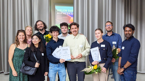 Group photo of around twelve people standing indoors. The winners from Hochschule Wismar hold certificates and flowers while organisers and supporters smile alongside them.