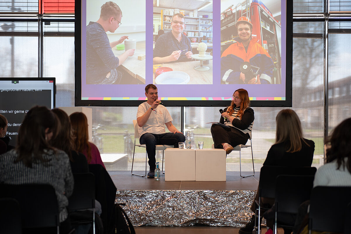 Merle Eva Pagel und Jannis Pagel stehen auf der Bühne und halten ihren Vortrag über Inklusion und persönliche Alltagserfahrungen. Das Publikum ist von hinten zu sehen.