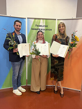 Die drei Personen stehen in einem Raum vor drei farbigen Bannern auf denen der Schriftzug "Feierliche Immatrikualtion 2025" steht.