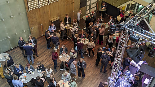 Es sind aus einer oberen Etage herab runde Stehtische an einem Lichthof zu sehen. An denen stehen Menschen und unterhalten sich.