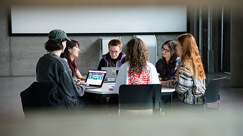 Sechs Personen sitzen im Kreis an einem sechseckigen Tisch. Auf ihren Laptops sind Grafik-Entwürfe zu sehen.