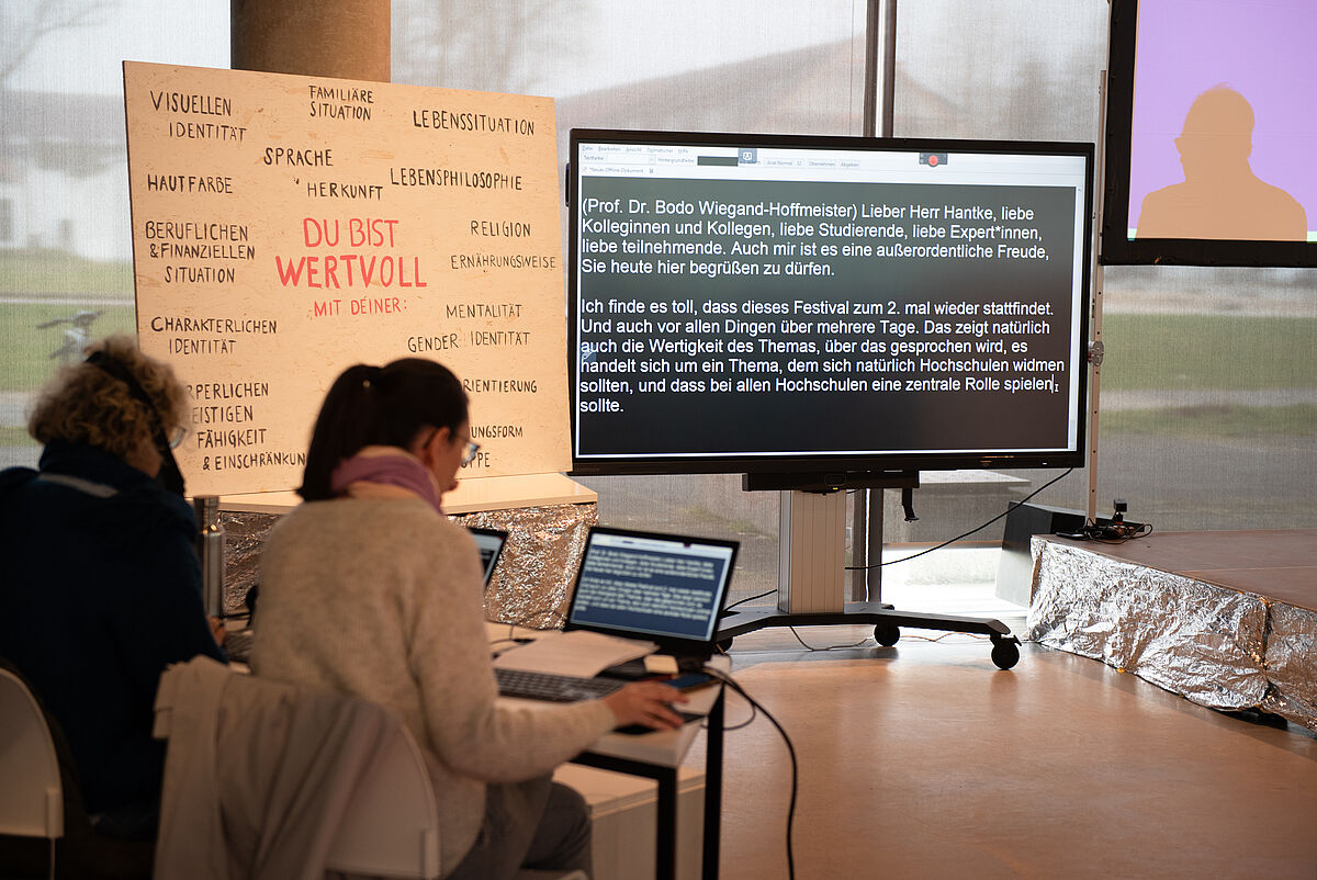 Zwei Schriftdolmetscherinnen sitzen mit dem Rücken zur Kamera und blicken auf ihre Laptops. Vor ihnen steht ein großer Bildschirm neben der Bühne, auf dem live gesprochene Inhalte für hörgeschädigte Personen verschriftlicht angezeigt werden.