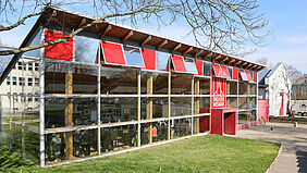 Theater Es ist das Theaterfoyer von außen zu sehen. Hinter den großen Glasscheiben sind viele Menschen, die sich unterhalten zu sehen.