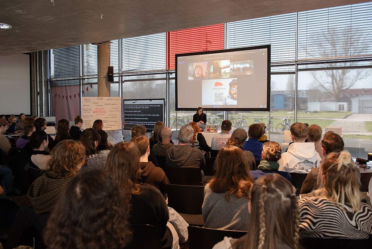 Blick von hinten über das Publikum, welches den Vortrag von Raúl Krauthausen auf einer großen Leinwand auf der Bühne verfolgt.