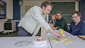 Ein Student steht links von einem Tisch und zeigt auf eine gelbe Skulptur, die einer Wachskerze ähnelt. Rechts vom Tisch sitzen die beiden Professoren und schauen direkt auf die Skulptur.