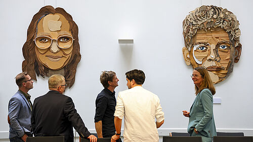 An einer Wand hängen zwei überlebensgroße Porträts einer weiblichenund einerr männlichen PErsonaus verschiedenfarbigen Holzteilen. Davor stehen die genannten Personen und unterhalten sich.