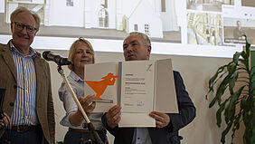 Gruppe Die drei Gratulanten stehen vor einer Leinwand, auf der die Preisträgerin online zu sehen ist.