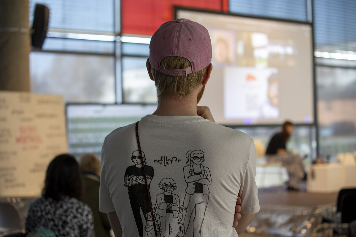 Ein Teammitglied steht mit dem Rücken zur Kamera im Veranstaltungsraum. Auf seinem T-Shirt ist die Festivalillustration des reflektor – Festival der Kommunikation abgebildet. Vor ihm ist auf der Bühne eine große Leinwand zu sehen, auf der Raul Krauthausen digital zugeschaltet ist und seinen Vortrag hält.