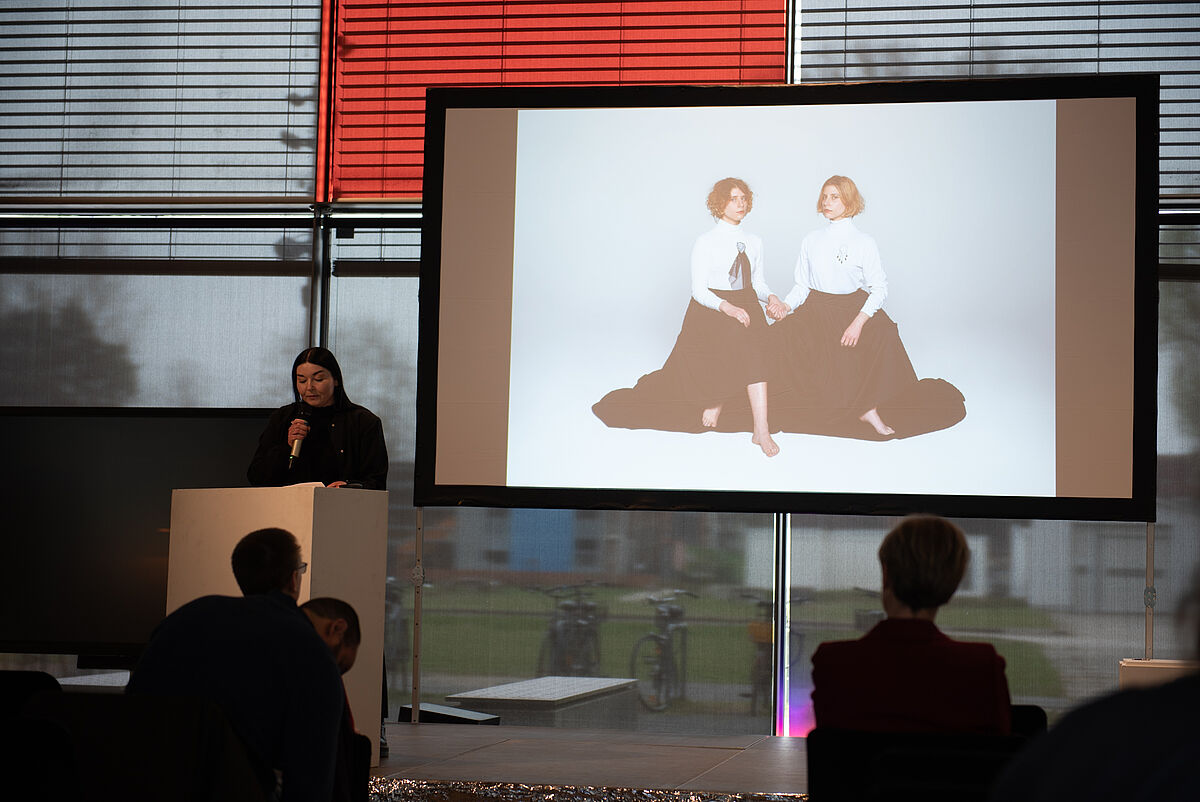 Sarina Drescher steht auf der Bühne und spricht über ihr Projekt „Cor“. Hinter ihr ist auf einem großen Bildschirm ein Bild zum Projekt zu sehen.