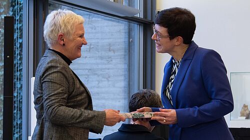 Prof. Polkehn hat sich von ihrem Stuhl erhoben und nimmt das Geschenk entgegen. Die um sie herum sitzenden Teilnehmenden schauen zu.