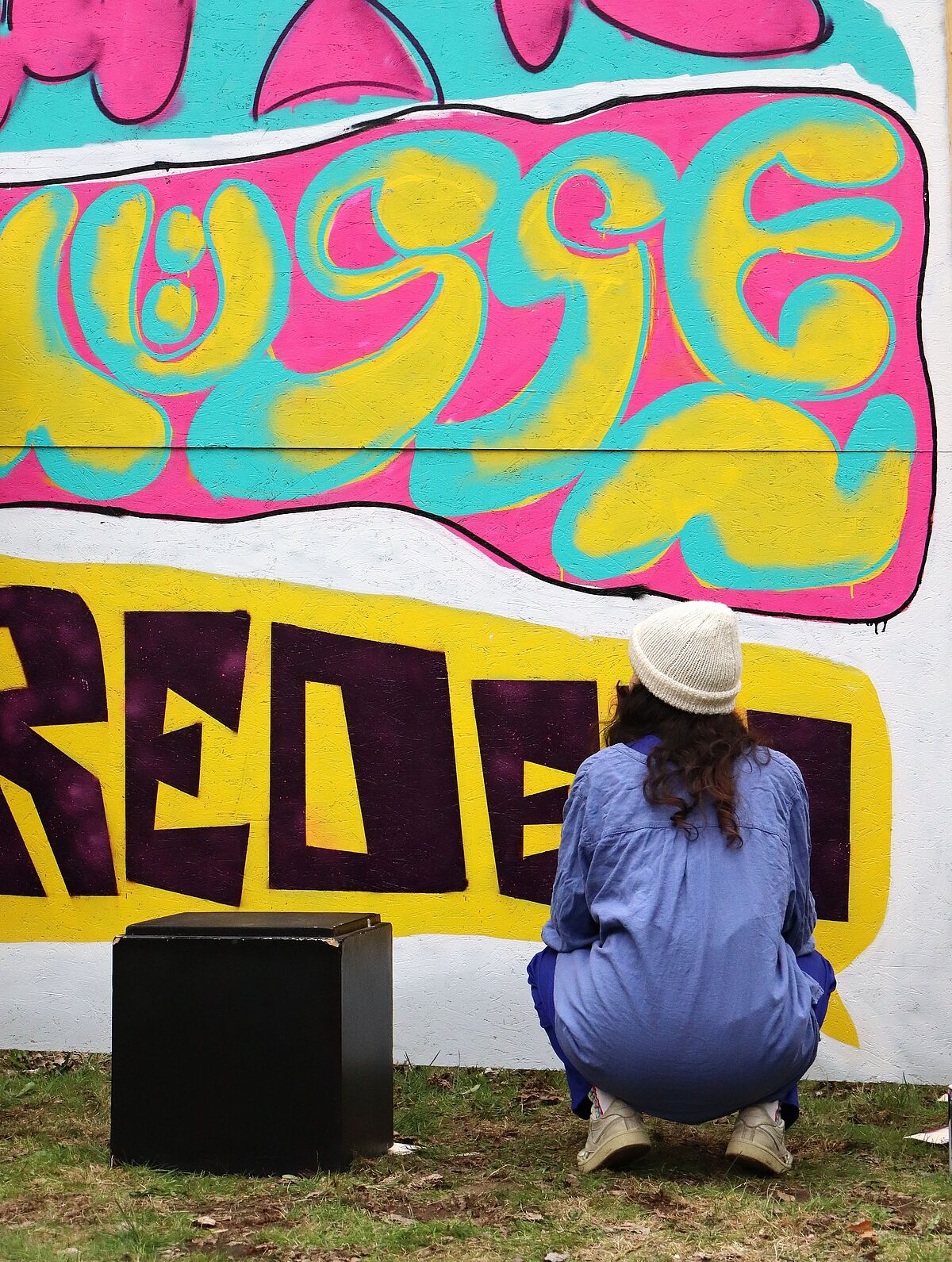 Eine Teilnehmerin hockt vor einer großen besprayten Wand und schaut auf ihr Werk während des Graffiti-Workshops.