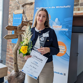 Female student Svea Bublys holds her winner certificate, trophy and a bouquet of yellow and white flowers. She stands in front of an inspired competition banner and a wooden signpost.