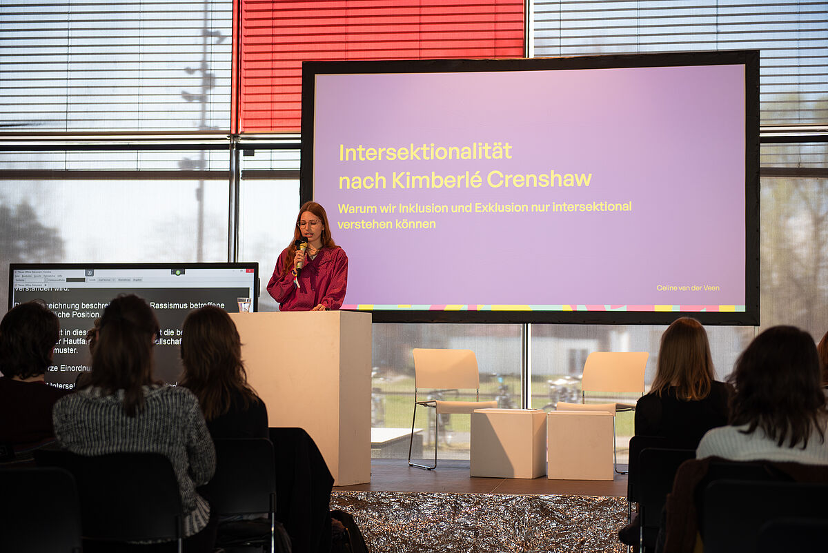 Celine van der Veen steht auf der Bühne und hält ihren Vortrag über Intersektionalität. Das Publikum ist von hinten zu sehen.