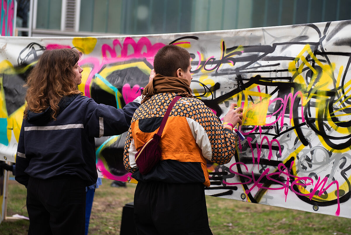 Zwei Personen sprühen gemeinsam Farbe auf ein Banner im Graffiti-Workshop, der sich künstlerisch mit den Themen Inklusion und Exklusion auseinandersetzt.