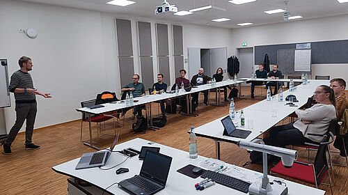 Ralmo Bode stands at the front of the seminar room and presents his research work. A slide with diagrams can be seen in the background, and the participants listen attentively.