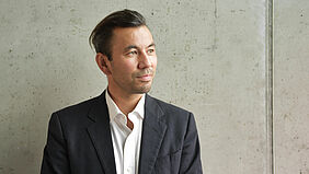 Takasaki-Lauw Es ist ein Porträt Prof. Takasaki-Lauws zu sehen. Dabei hat steht er vor einer Sichtbetonwand im Haus 7.