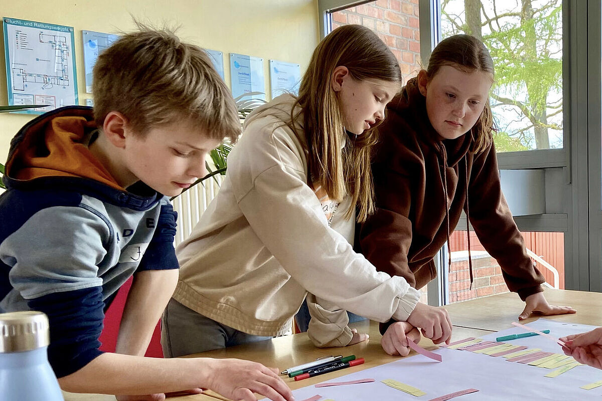 Drei Kinder stehen an einem Tisch und schieben beschriebene Papierschnipsel