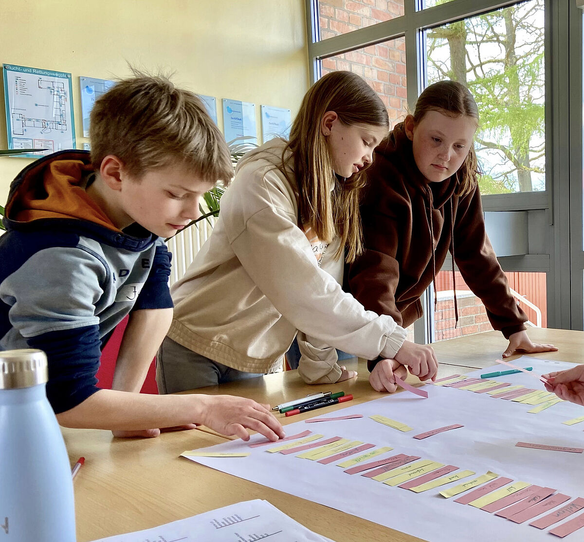 Drei Kinder stehen an einem Tisch und schieben beschriebene Papierschnipsel