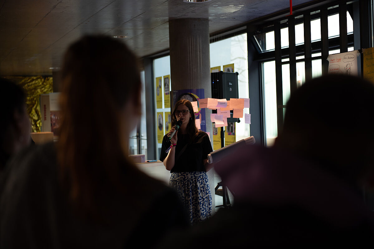 Sara steht vor dem Publikum und eröffnet die Postersession, bei der die Teilnehmenden Einblicke in die Ergebnisse der verschiedenen Workshops erhalten.
