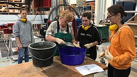 im Labor Fünf Studentinnen stehen um eine Frau herum - alle haben Gummihandschuhe an. Vor der Frau stehen zwei Eimer auf einem Tisch. Aus einem der Eimer entnimmt sie eine Michschung mit eienr Schaufel.