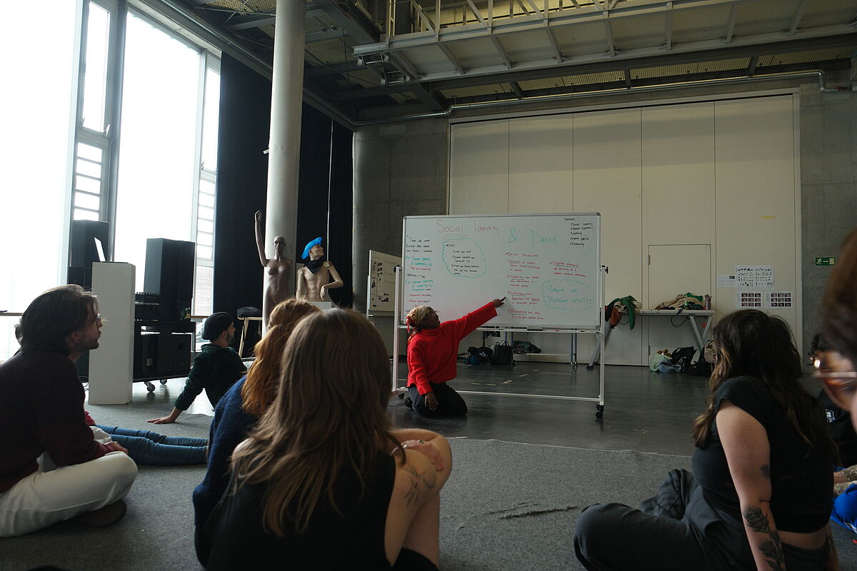Accacia sitzt zusammen mit den Teilnehmenden auf dem Boden im Workshop "Social Identity and Dance" und zeigt auf eine Tafel, um etwas zu erklären.