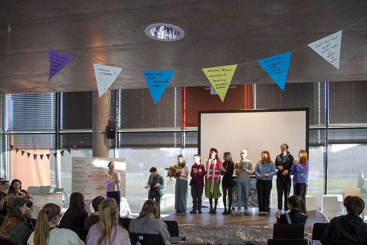 Das studentische Team von Reflektor steht auf der Bühne. Sara, die Projektleiterin, erhält einen Blumenstrauß als Anerkennung für ihr Engagement.