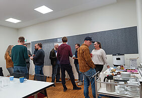 Participants stand together in small groups, talking to each other and holding cups of punch. Plates of pastries are on the tables.