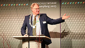 Rektor Es ist der Rektor als Redner hinter dem Pult und vor Banner mit dem Aufdruck „Jahresempfang 2023“ zu sehen, wie er mit ausgebreitetem Arm freundlich lächelnd zur Seite schaut.