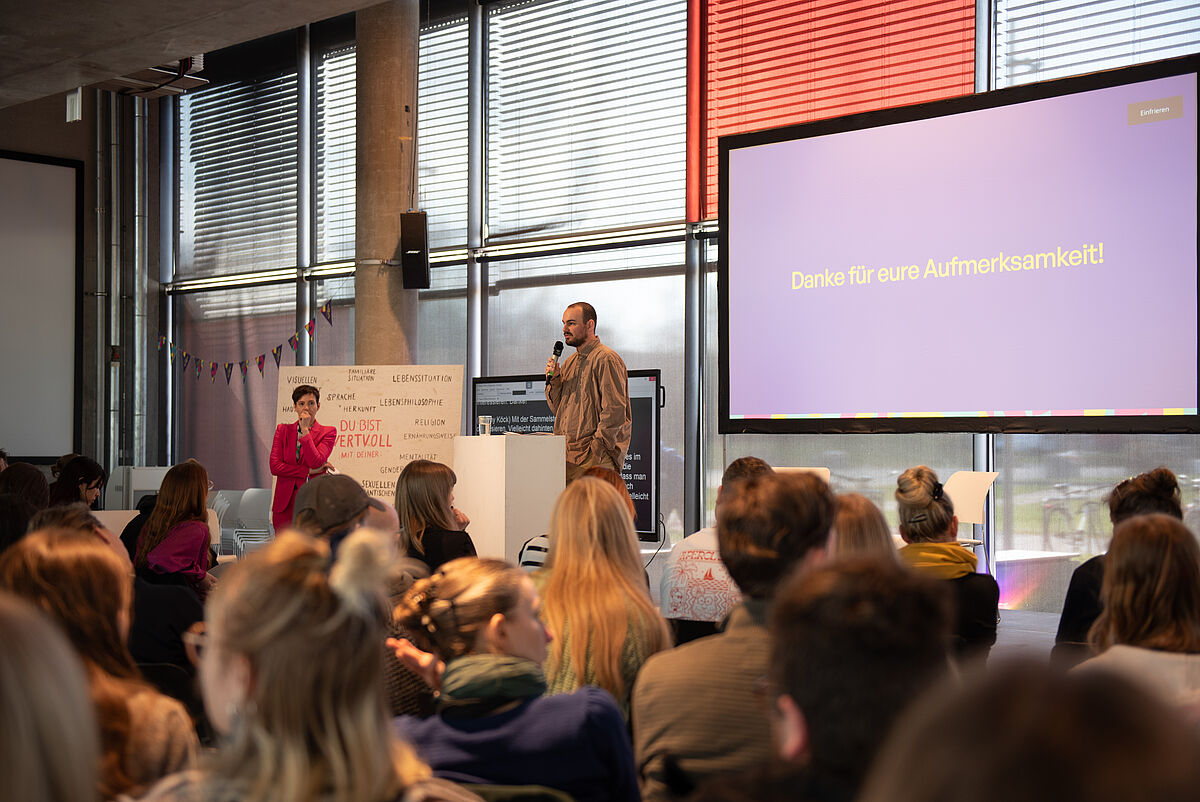 Jeremy Köck steht auf der Bühne und hält einen Vortrag zum Thema Inklusion im Studium. Das Publikum ist von hinten zu sehen.