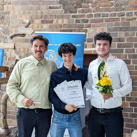 Three male students from the Navoko team stand in front of a brick wall. They hold winner certificates and a trophy while smiling confidently.