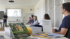Der Blick geht über Broschüren und an einem Tisch sitzende junge Menschen nach vorn zu einem Livebild. Darauf ist eine Teilnehmerin zu sehen, die live zugeschaltet ist. Davor steht Jan Oestreich.