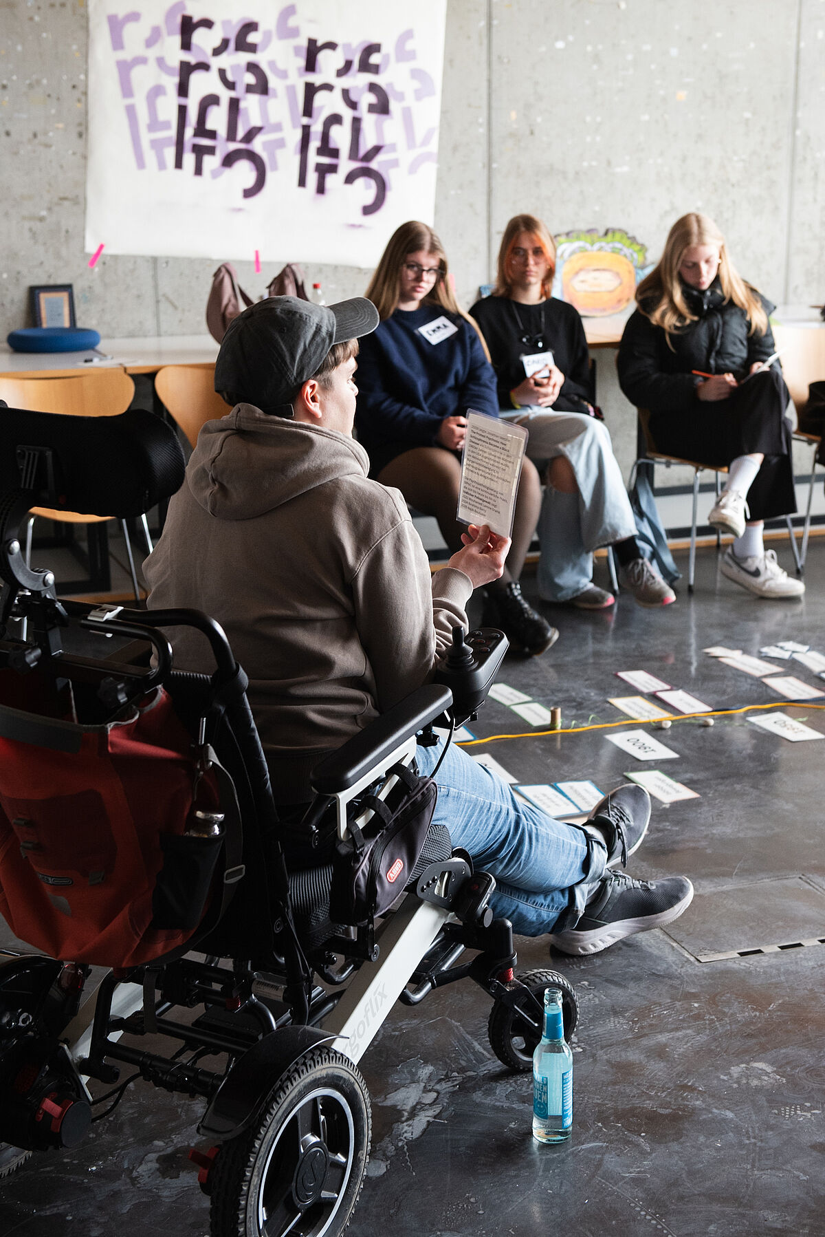 Eine Person im Rollstuhl hält eine Karte in der Hand und spricht, um sie herum sitzen Teilnehmende auf dem Boden, auf dem weitere Karten ausgelegt sind.