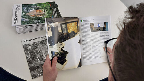 You look over the shoulder of a person holding an unfolded brochure in their hands. On the table are more copies and an insert that is slightly smaller than the brochure printed in A4 format.