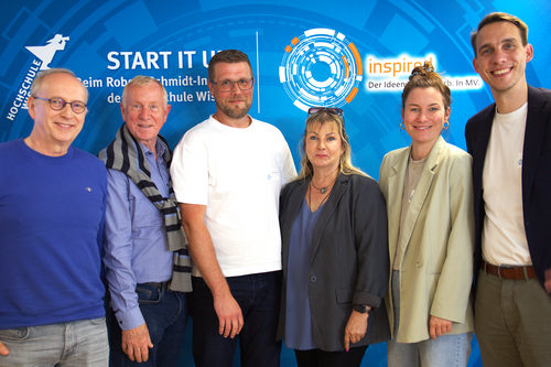 Group photo of six jury members of the INSPIRED competition standing in front of a branded background at Wismar University of Applied Sciences.