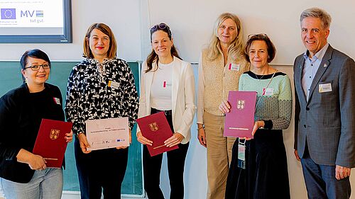 Five women and one man are standing next to each other. They are holding document folders with the state government's logo. In the background, a presentation with the name of the Minister of Justice can be seen.