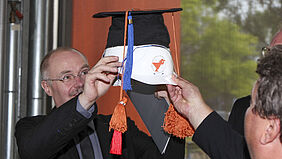 Kathmann hält einen weißen Helm auf dem sih ein schwarzer Doktorhut befindet, in Kopfhöhe, um ihn aufzusetzen.