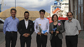 Gruppe Die fünf Personen stehen nebeneinander. Im Hintergrund ist ein Schiff, ein Kran und ein Kieshaufen im Wismarer Seehafen zu erkennen.