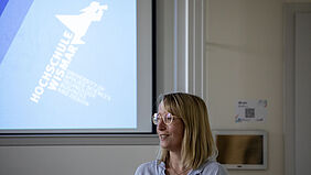 A young woman wearing glasses and a striped blouse stands in front of the projection screen for her slide presentation.