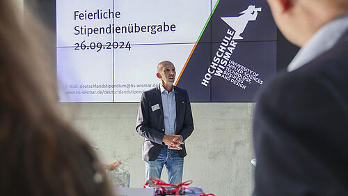 Der Fotograf schaut über die Schulter zweier Stipendiaten. Der Prorektor steht vor einer Bildschirmpräsentation auf der Datum und Veranstaltungstitel stehen.