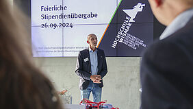 Der Fotograf schaut über die Schulter zweier Stipendiaten. Der Prorektor steht vor einer Bildschirmpräsentation auf der Datum und Veranstaltungstitel stehen.