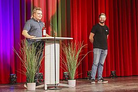 AStA-Vorsitz Lukas steht hinter dem Rednerpult auf der Bühne, während vor ihm eine Flashce mit roter Flüssigkeit steht. NEben ihm verschränkt eine weitere PErson ihre Armen hinter dem Rücken und schaut lachend zu Lukas hinüber.