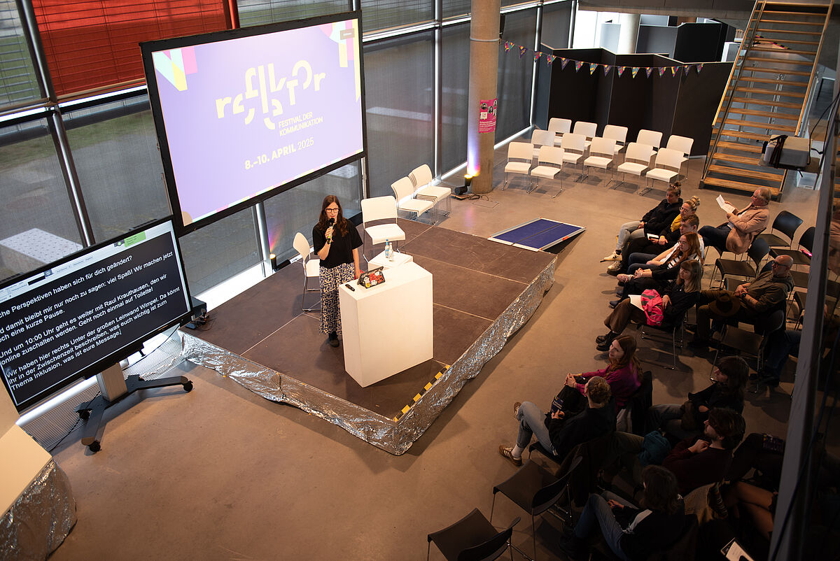 Sara Scholl, studentische Projektleiterin des reflektor Festivals, steht auf der Bühne und spricht in ein Mikrofon. Sie richtet zur Eröffnung des Festivals ein Grußwort an das Publikum.